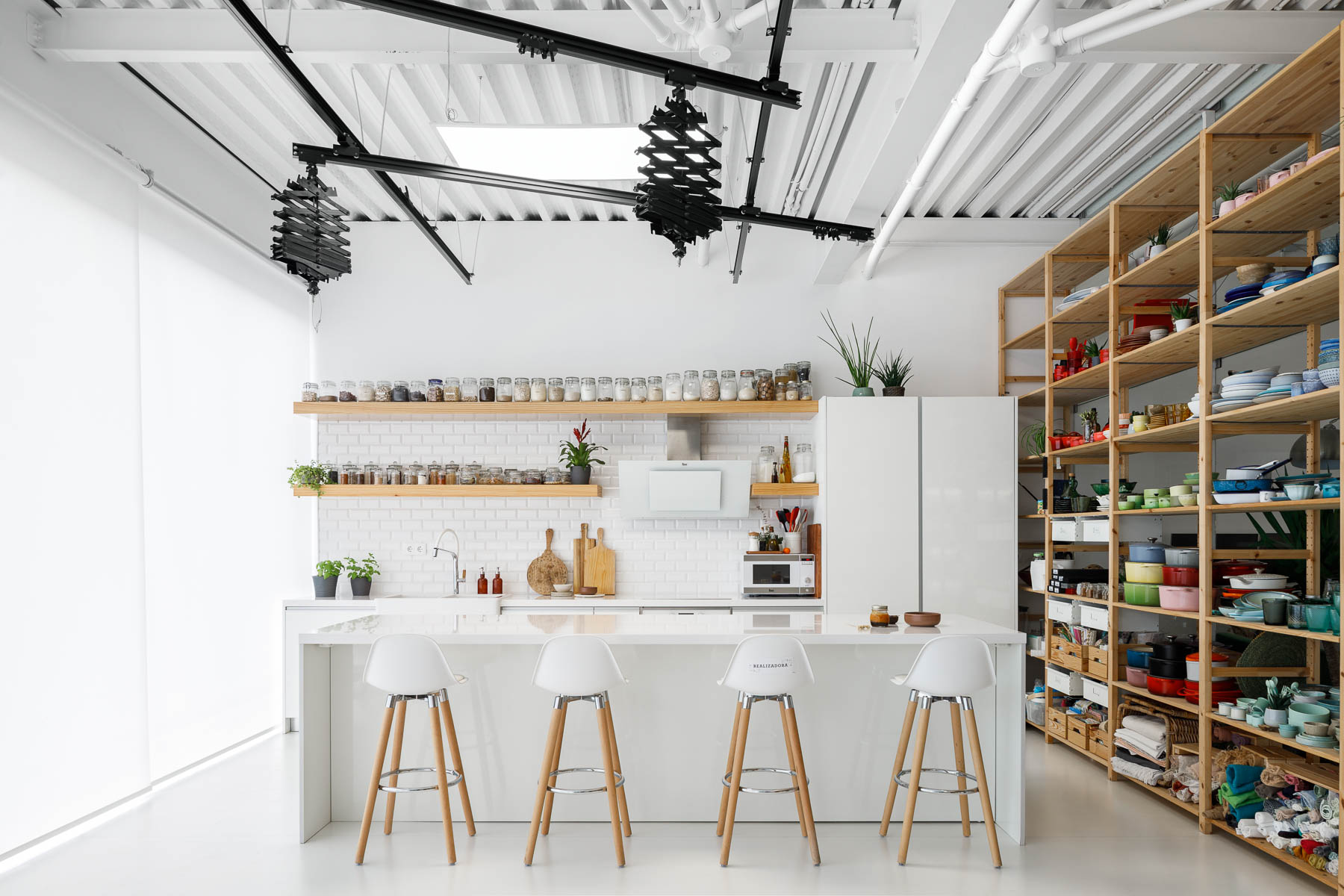 A modern office has a photo studio with a minimalist white kitchen with floating wood shelves. Adjacent to the kitchen are tall wood shelves filled with photography props and kitchen equipment.